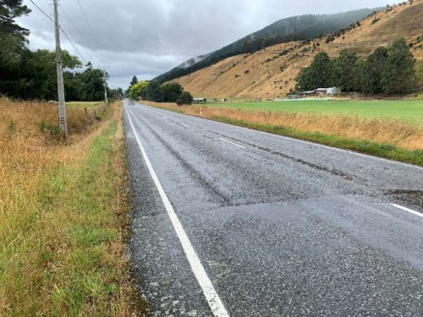 A wet, empty road stretches into the distance between grassy fields and rolling hills under a cloudy sky, creating a calm, rural landscape.