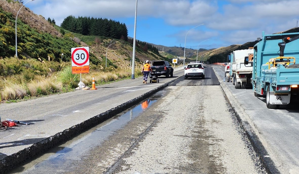 A rural road under construction with a temporary 30 km/h speed limit sign. Cars drive past workers and construction vehicles. The sky is partly cloudy.